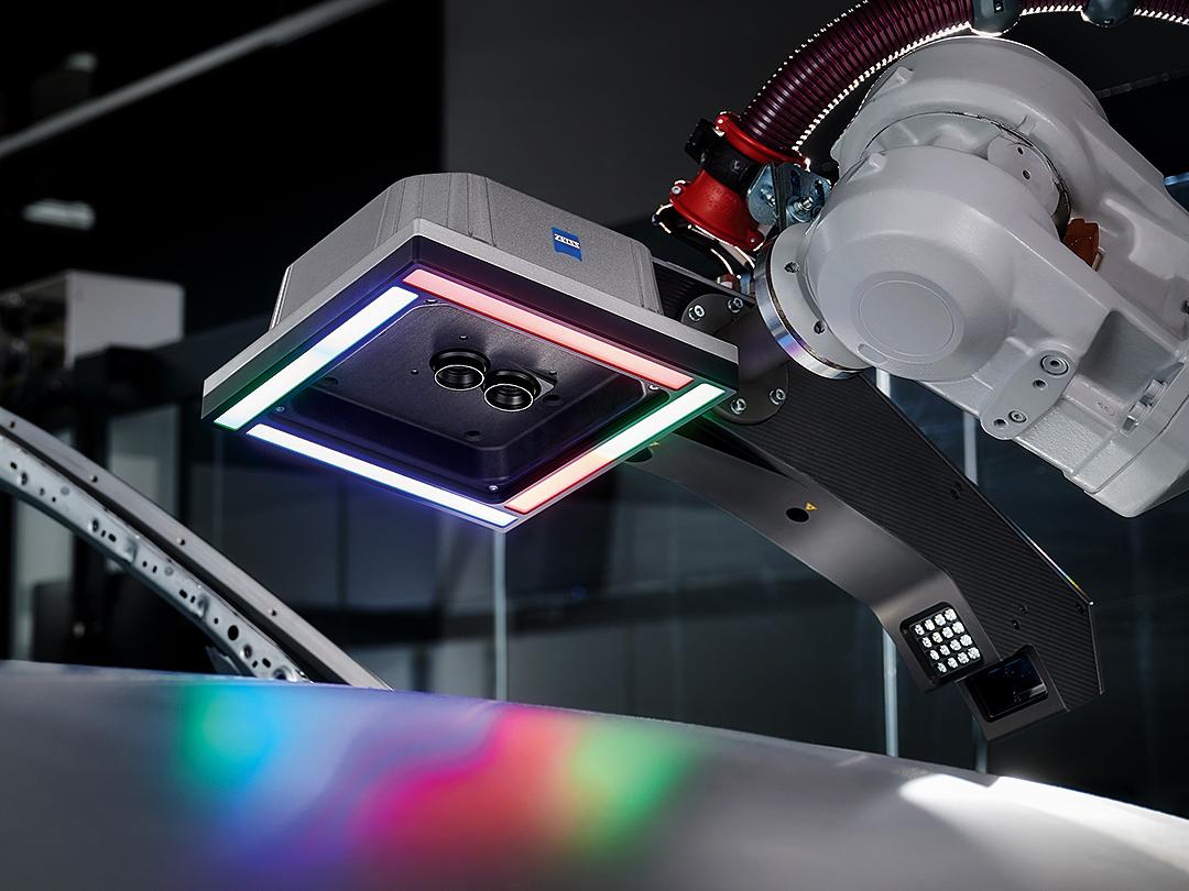 Một cánh tay robot được trang bị cảm biến quét 3D ZEISS lơ lửng phía trên bề mặt kim loại, được chiếu sáng bằng đèn LED nhiều màu.